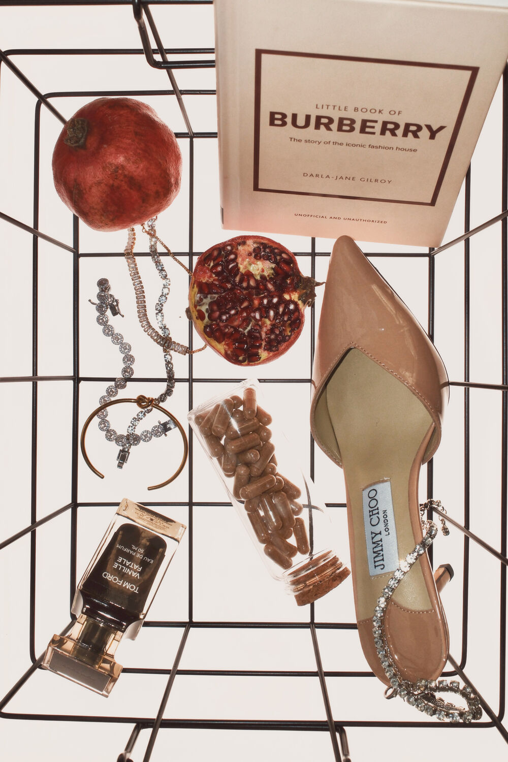 Still life in a wire basket: Burberry book, pomegranate, Jimmy Choo heel, Tom Ford perfume